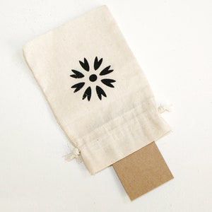 May include: A small white cotton drawstring bag with a black flower design. The bag is open and a brown card is partially visible inside.