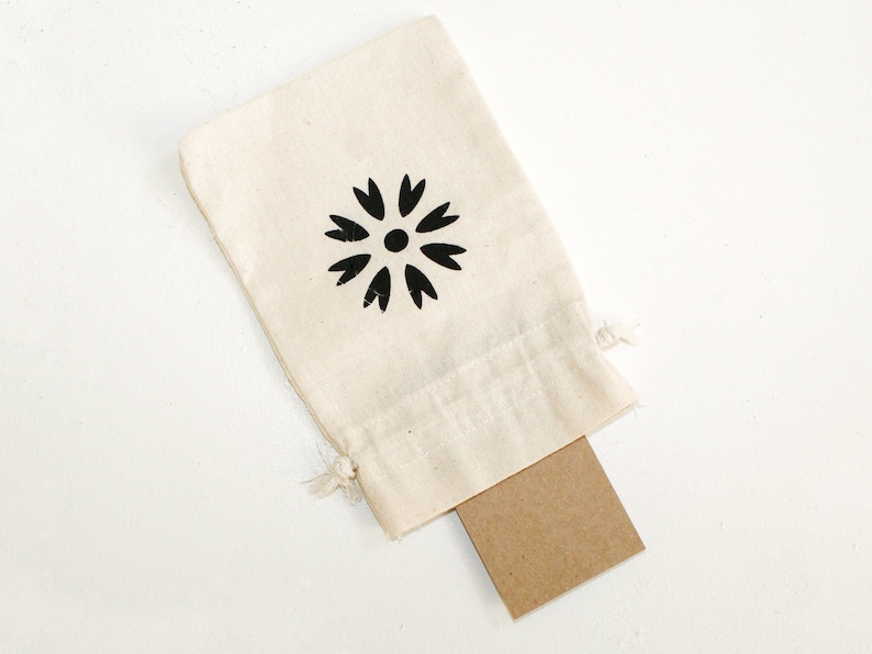 May include: A small white cotton drawstring bag with a black flower design. The bag is open and a brown card is partially visible inside.