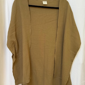 May include: An olive green open-front kimono-style garment made of a textured, crinkled fabric. The piece features wide, short sleeves and a loose, flowing silhouette. A small rectangular brand label is visible at the inside of the neckline.