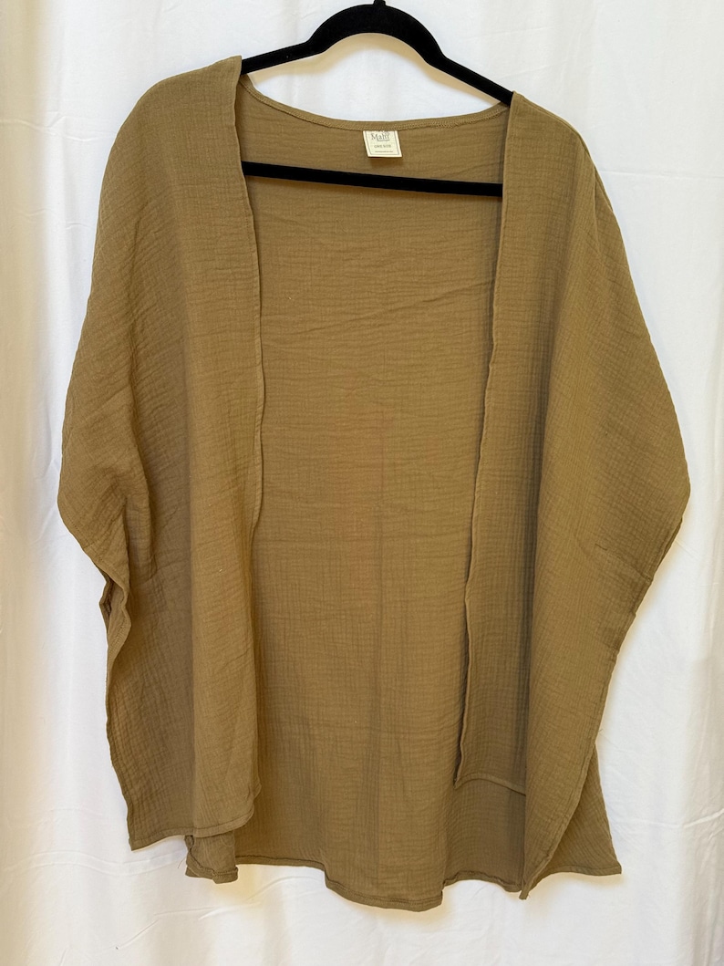 May include: An olive green open-front kimono-style garment made of a textured, crinkled fabric. The piece features wide, short sleeves and a loose, flowing silhouette. A small rectangular brand label is visible at the inside of the neckline.