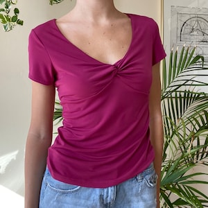 Puede incluir: Una camiseta magenta de manga corta con un detalle retorcido en el escote.