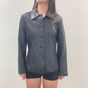 May include: A black leather jacket with a collared neckline and button-down closure. The jacket has long sleeves and a tailored fit. The jacket is worn with black shorts. The jacket appears to be a vintage or pre-owned item.