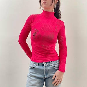 Pode incluir: Uma blusa rosa brilhante de manga comprida com gola alta e com babados. A blusa apresenta um design de malha aberta texturizada com detalhes recortados nas mangas e no peito. A pe&ccedil;a &eacute; combinada com jeans azuis claros.