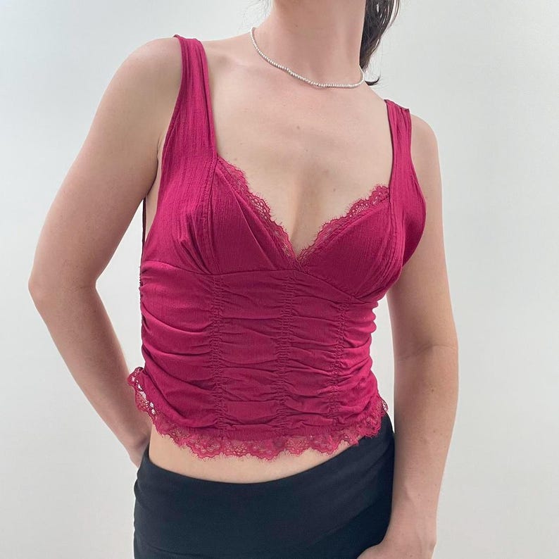 May include: A magenta-colored camisole top with a V-neckline and delicate lace trim. The top features a ruched bodice and thin straps. This stylish top is a great addition to any wardrobe.