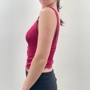 May include: A sleeveless, deep magenta top with a V-neckline and delicate lace trim. The fabric is ruched along the sides, adding texture to the garment. The top is a solid color and appears to be made of a lightweight material.