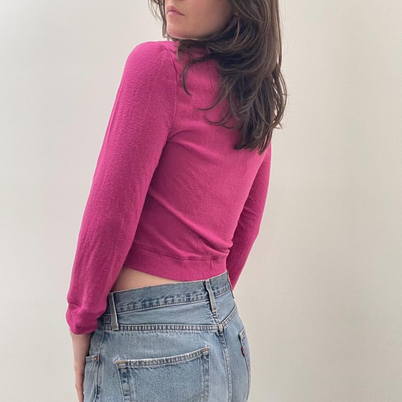 May include: A pink long-sleeved cropped sweater with a ribbed hem. The sweater is worn with blue jeans.