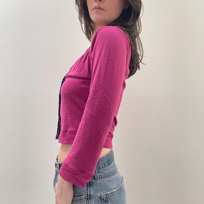 May include: A magenta long-sleeved cardigan with a black trim. The cardigan is cropped and has a slightly loose fit.