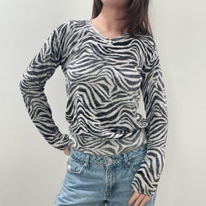 May include: A long-sleeved top with a zebra print in black and white. The top has a V-neck and is paired with light blue denim jeans. The zebra pattern covers the entire top, creating a bold, eye-catching design.
