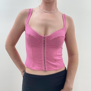 Puede incluir: Un top bustier rosa acanalado con escote corazón y cierre de cremallera metálica en la parte delantera. El top tiene tirantes anchos y una silueta ajustada. La prenda es de color rosa liso.