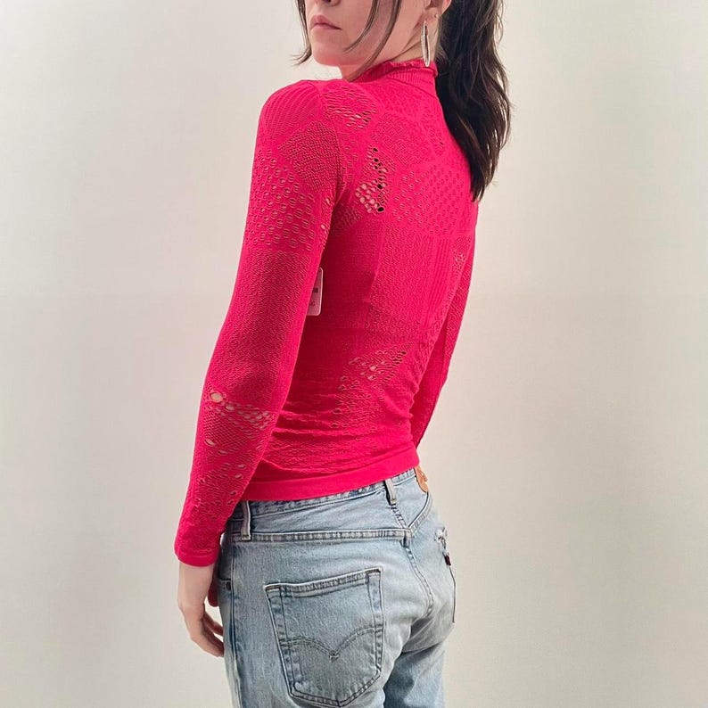 Pode incluir: Uma blusa rosa brilhante de manga comprida com gola alta. A blusa tem um design de malha aberta texturizada com detalhes recortados. A modelo est&aacute; usando jeans azuis claros. A blusa tem uma etiqueta.