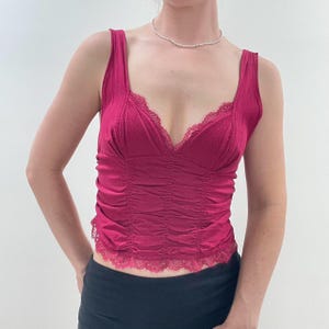 May include: A magenta-colored tank top with a V-neckline and lace trim. The top features a ruched design on the front and wide straps. The garment is made of a textured fabric and is a stylish piece of clothing.