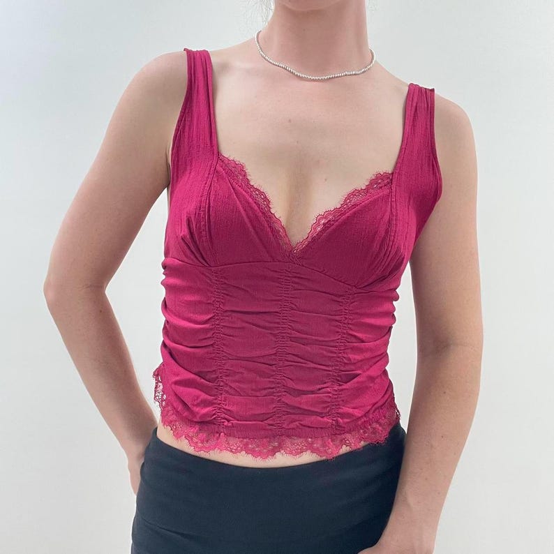May include: A magenta-colored tank top with a V-neckline and lace trim. The top features a ruched design on the front and wide straps. The garment is made of a textured fabric and is a stylish piece of clothing.