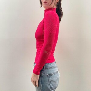 Pode incluir: Uma blusa rosa brilhante de manga comprida com gola alta. A blusa tem um padr&atilde;o texturizado e detalhes recortados nas mangas. A modelo est&aacute; usando jeans azul claro. A blusa &eacute; justa e feita de um material el&aacute;stico.