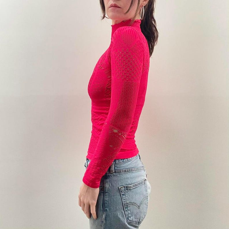 Pode incluir: Uma blusa rosa brilhante de manga comprida com gola alta. A blusa tem um padr&atilde;o texturizado e detalhes recortados nas mangas. A modelo est&aacute; usando jeans azul claro. A blusa &eacute; justa e feita de um material el&aacute;stico.