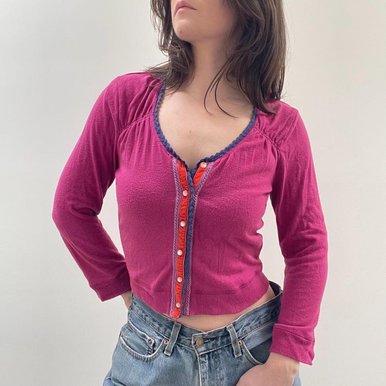 May include: A pink long-sleeved cardigan with a button-up front and a blue and orange trim. The cardigan has a V-neck and is cropped.