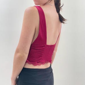 May include: A burgundy tank top with a low back and thin straps. The top features a ruched design on the front and a lace trim along the bottom edge. The model is wearing black pants.