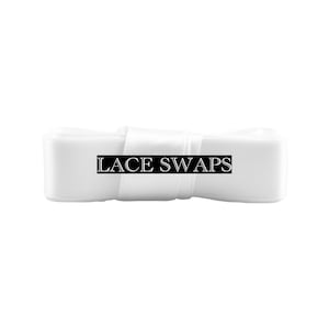 May include: A roll of smooth white satin ribbon featuring a black rectangular label centered on the front. The label contains the white text LACE SWAPS in a serif font. The ribbon is tightly wound and has a consistent, clean appearance.
