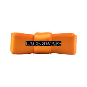 May include: A bright orange satin ribbon folded into a rectangular shape with a central band. The text LACE SWAPS is printed in a white serif font on a black rectangular background centered across the front of the orange ribbon.