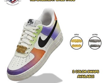 May include: A pair of gold-colored metal grillz-style lace tags attached to the laces of a white sneaker with a multi-colored gradient pattern. The image displays gold and silver options for these replacement lace tags, labeled as Grillz Metal Dubrae.