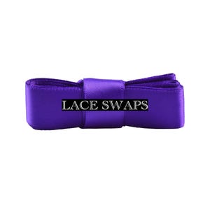 May include: A vibrant purple satin ribbon folded into a compact rectangular shape, featuring a smooth, reflective texture. The text LACE SWAPS is printed in a clear, white serif font across the center of the ribbon, which is secured by a matching purple band.