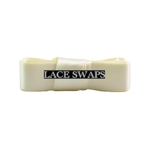 May include: A folded length of cream-colored satin ribbon with a smooth, reflective texture. A black rectangular label is centered on the ribbon, featuring the white text LACE SWAPS in a serif font. The ribbon is neatly coiled and layered.