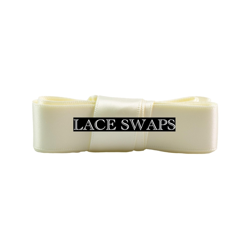 May include: A folded length of cream-colored satin ribbon with a smooth, reflective texture. A black rectangular label is centered on the ribbon, featuring the white text LACE SWAPS in a serif font. The ribbon is neatly coiled and layered.