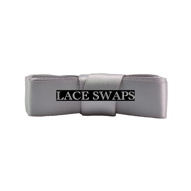 May include: A light gray satin ribbon folded into a rectangular shape with a central band. The text LACE SWAPS is printed in white capital letters on a black rectangular background centered across the front of the smooth, reflective fabric.