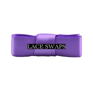 May include: A vibrant purple satin ribbon folded into a compact bundle, featuring a smooth, reflective texture. A black rectangular label with the white text LACE SWAPS is centered horizontally across the front of the ribbon material.
