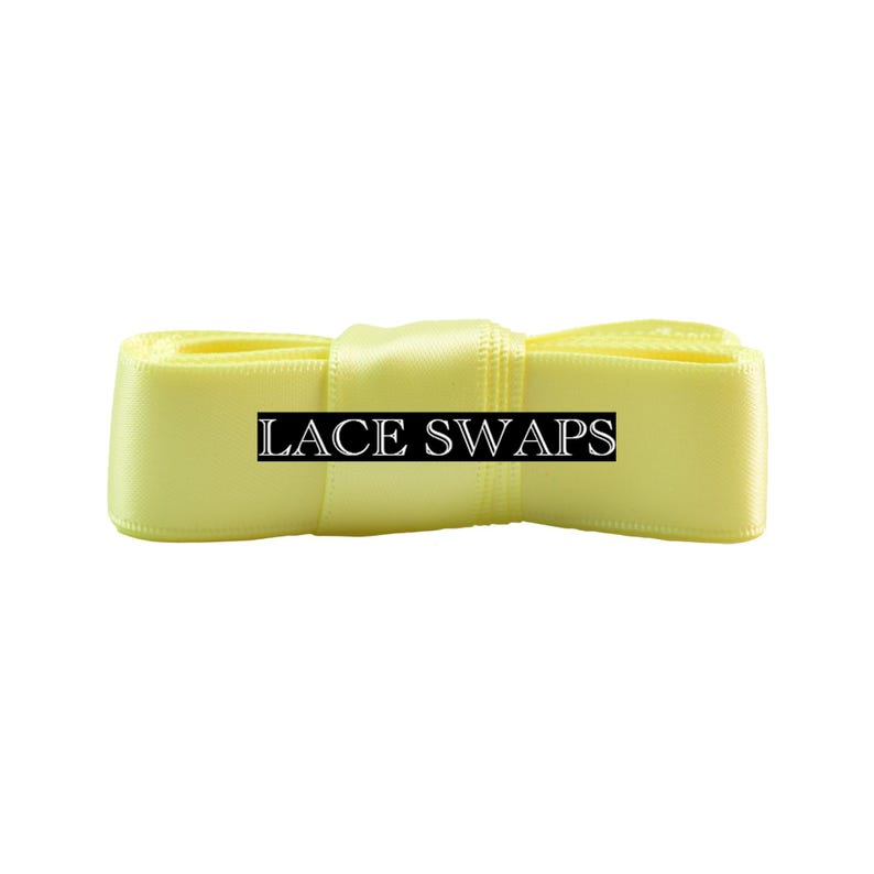 May include: A pale yellow satin ribbon folded into a bow shape. The center of the ribbon features a black rectangular label with the white text LACE SWAPS printed in a serif font. The ribbon has a smooth, reflective texture and a consistent color.