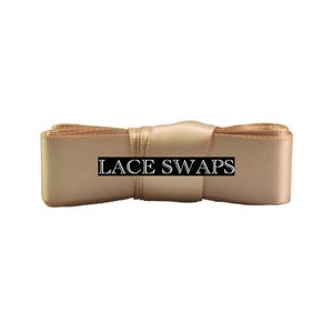 May include: A roll of champagne-colored satin ribbon featuring a smooth, reflective texture. A black rectangular label with the white text LACE SWAPS is centered horizontally across the front of the ribbon, which is coiled into a flat bundle.