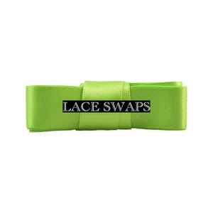 May include: A bright lime green satin ribbon folded into a rectangular shape. A black rectangular label is centered on the ribbon with the white text LACE SWAPS printed in a serif font. The ribbon has a smooth, reflective texture and consistent color.