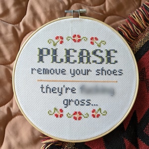Pode incluir: Um aro de bordado branco com um desenho de ponto de cruz que diz "PLEASE remove your shoes they're gross..." com flores vermelhas ao redor do texto.