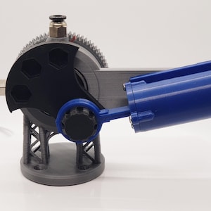 May include: A mechanical device with a gear, a blue cylinder, and a black hexagonal plate. The device is gray and black, with a blue cylinder attached. The gear has teeth around the edge. The device is on a white surface.