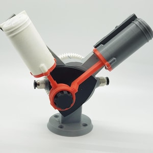 May include: A 3D-printed model of a mechanical device, featuring a V-shaped design with two cylindrical components in white and gray. The model incorporates orange and black accents, a circular base, and a gear mechanism.