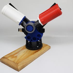 May include: A 3D-printed model featuring two angled cylinders, one white and one red, connected by a blue and black mechanism. The model is mounted on a wooden base, showcasing a combination of geometric and organic shapes.