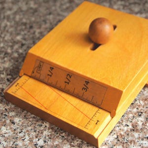 Puede incluir: Una herramienta de madera con una regla deslizante, con medidas en fracciones. La herramienta tiene una pequeña bola de madera en la parte superior y una línea roja dibujada en la regla. La madera tiene un acabado claro y natural.