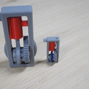 May include: Two gray 3D-printed models of an engine, one large and one small, sit on a light wood surface. The engines have red cylinders and blue and gray internal components. The gears and mechanical parts are visible.