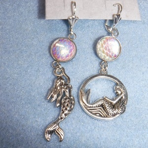 May include: A pair of silver mermaid earrings with iridescent pink and white cabochon accents. Each earring features a mermaid charm, one with a mermaid curled in a circle and the other with a mermaid swimming.