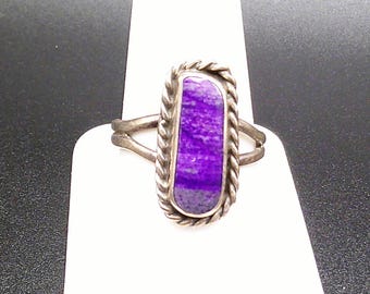 May include: A silver ring with a rectangular, vivid purple stone. The stone is framed by a twisted rope-style border. The ring's band splits into two delicate arms either side of the stone.