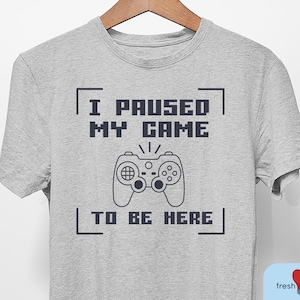 May include: A gray t-shirt with a black graphic that says "I paused my game to be here" with a pixelated game controller in the middle.