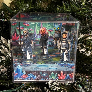 May include: A clear acrylic cube with a 3D pop-up scene featuring four characters. The scene is set in a colorful, fantastical landscape with glowing crystals and mushrooms. The characters include a bearded man in a black and green outfit, a woman in a black and red outfit, a blue creature with a pointed head, and a person in a white spacesuit.