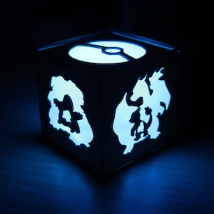 May include: A blue light illuminates a white cube with cut-out designs of Pokemon characters. The cube has a Pokeball design on the top.