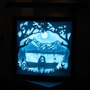 May include: A blue-lit, layered paper art piece depicting a romantic scene. A couple in a boat on a lake is silhouetted against a mountain backdrop, with trees and a full moon above. The artwork is framed in a black box.