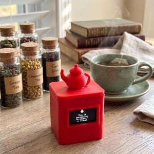 Puede incluir: Una caja cuadrada de color rojo brillante con una pequeña figura de tetera en la parte superior y una pantalla digital que muestra el texto: "Tea is permission to pause." --ritual. El dispositivo está junto a frascos de té y una taza.