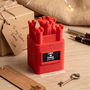 Pode incluir: Uma caixa vermelha em forma de castelo com um pequeno ecrã que mostra nomes e uma data. A caixa está ao lado de uma caixa de presente com uma etiqueta que diz "To my Queen Emma - Liam". Um marcador de madeira e uma chave decorativa também estão presentes.