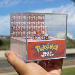 May include: A clear cube with a 3D diorama inside depicting a scene from the Pokemon Ruby video game. The diorama features a pixelated character standing on a brown and pink platform with a red background. The background features the Pokemon Ruby logo and the text "Ruby Version".