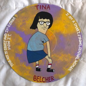 May include: A hand-painted vinyl record featuring a cartoon character with black hair, glasses, and a blue shirt. The character is standing with one leg bent and the other straight. The record is painted in shades of yellow, purple, and blue. The text "TINA BELCHER" is written in red at the bottom of the record. The text "I AM A SMART, STRONG, SENSUAL WOMAN" is written in white on the right side of the record. The text "YOUR ASS IS GRASS AND IM GONNA MOW IT." is written in white on the left side of the record.