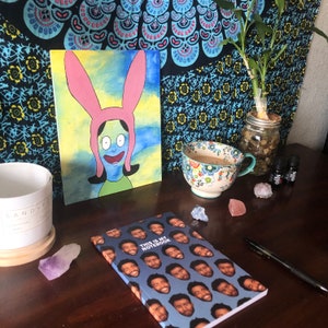 May include: A still life arrangement featuring a canvas painting of a cartoon character with pink bunny ears, a floral teacup, a notebook with a face pattern, and essential oil bottles. The notebook reads "This is my Notebook".