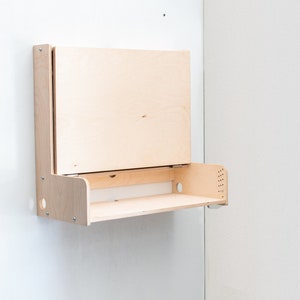 May include: A wall-mounted, fold-down desk made of light wood. The desk has a shelf and a hinged top that folds down to create a work surface.