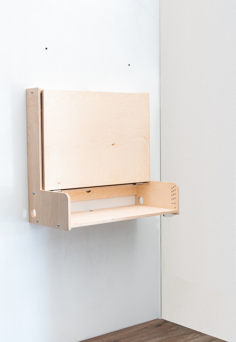 May include: A wall-mounted, fold-down desk made of light wood. The desk has a shelf and a hinged top that folds down to create a work surface.